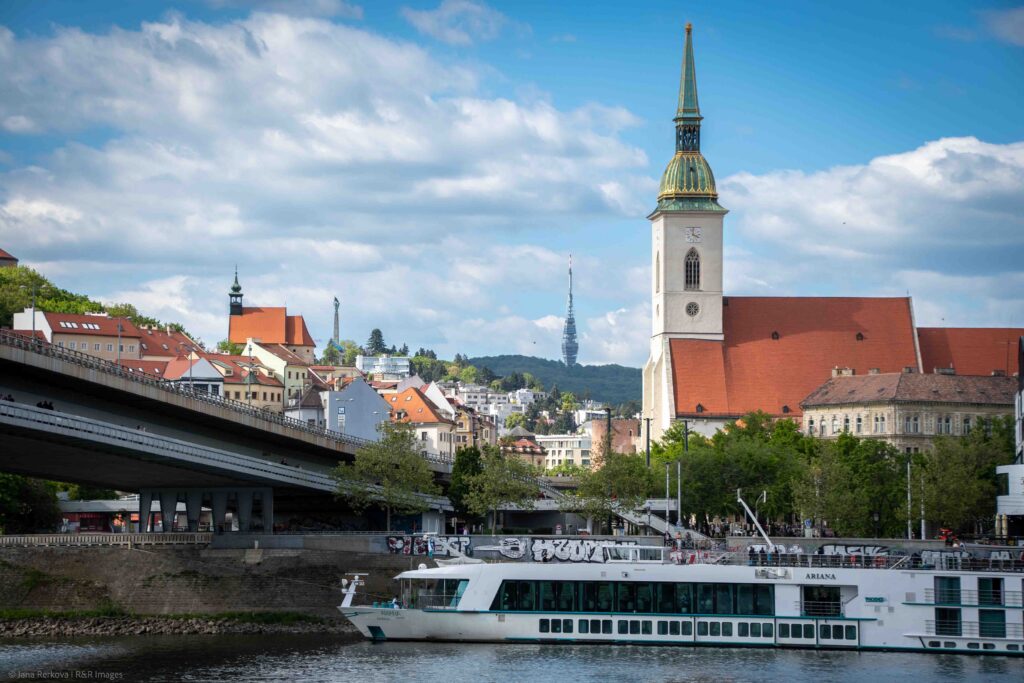 Bratislava cityscape from across the Danube, featuring the SNP Bridge, a river cruise, St. Martin's Cathedral, and Kamzík Tower.