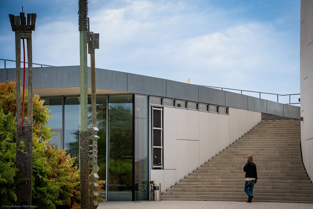 A modern architectural view of the exterior of the Danubiana Meulensteen Art Museum near Bratislava. A woman in a black jacket and jeans walks up a wide concrete staircase built alongside a curved, minimalist white building facade with floor-to-ceiling glass windows.