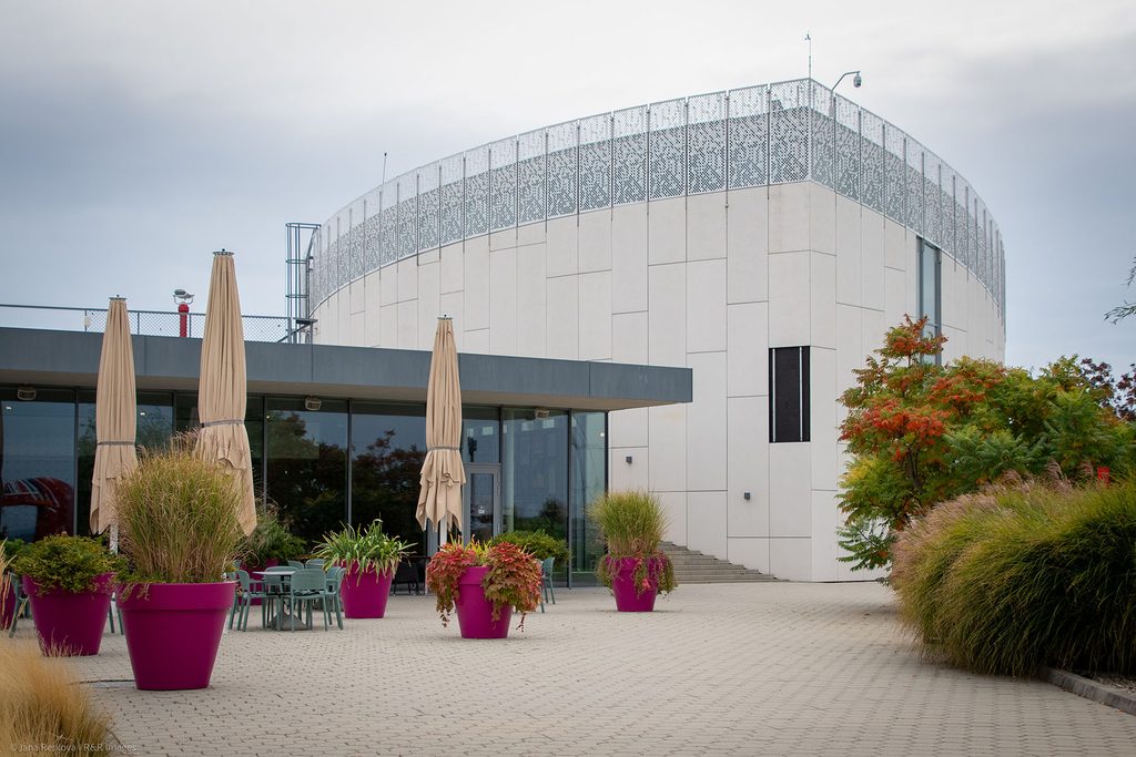 An exterior view of the modern Danubiana Meulensteen Art Museum, showcasing a curved white pavilion building, an outdoor paved terrace, cafe tables and chairs.