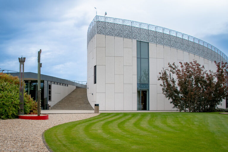 A wide exterior view of the contemporary, curved white building of the Danubiana Meulensteen Art Museum. A manicured green lawn with curved mowing patterns sweeps across the foreground next to a gravel path. Abstract outdoor sculptures and landscaping flank the modern structure, which features a wide staircase leading to an upper level under a cloudy sky.
