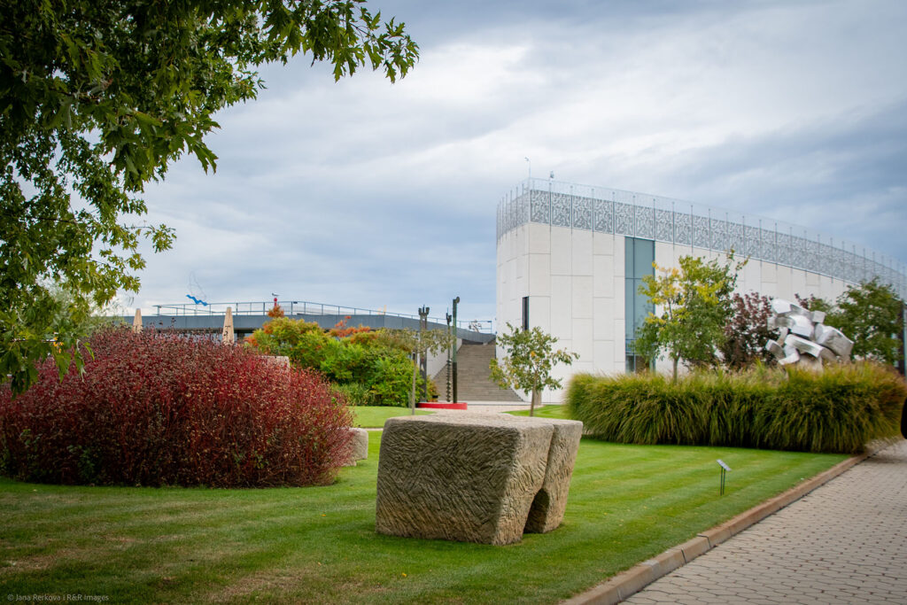 A scenic view of the Danubiana Meulensteen Art Museum grounds under a cloudy sky. In the foreground, a vibrant green lawn is accented by a dense red shrub, a chunky square stone sculpture, tall ornamental grasses, and scattered trees, all bordered by a paved walkway.