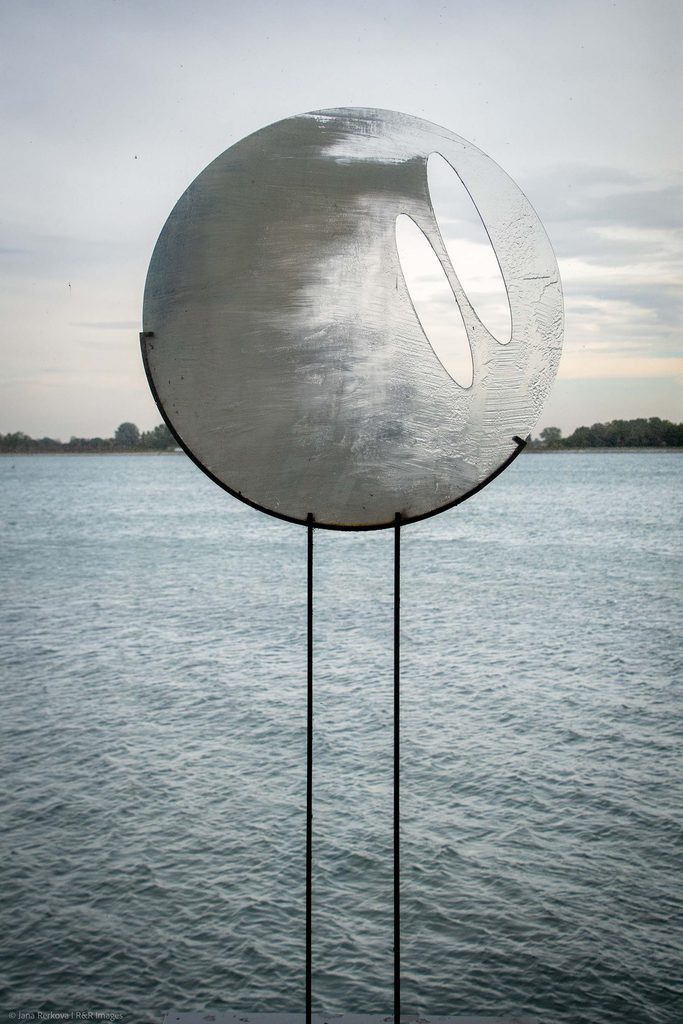 How Are You, Wind? by Palo Macho - A large metallic circular sculpture with two oval cutouts overlooking a wide river under a cloudy sky.
