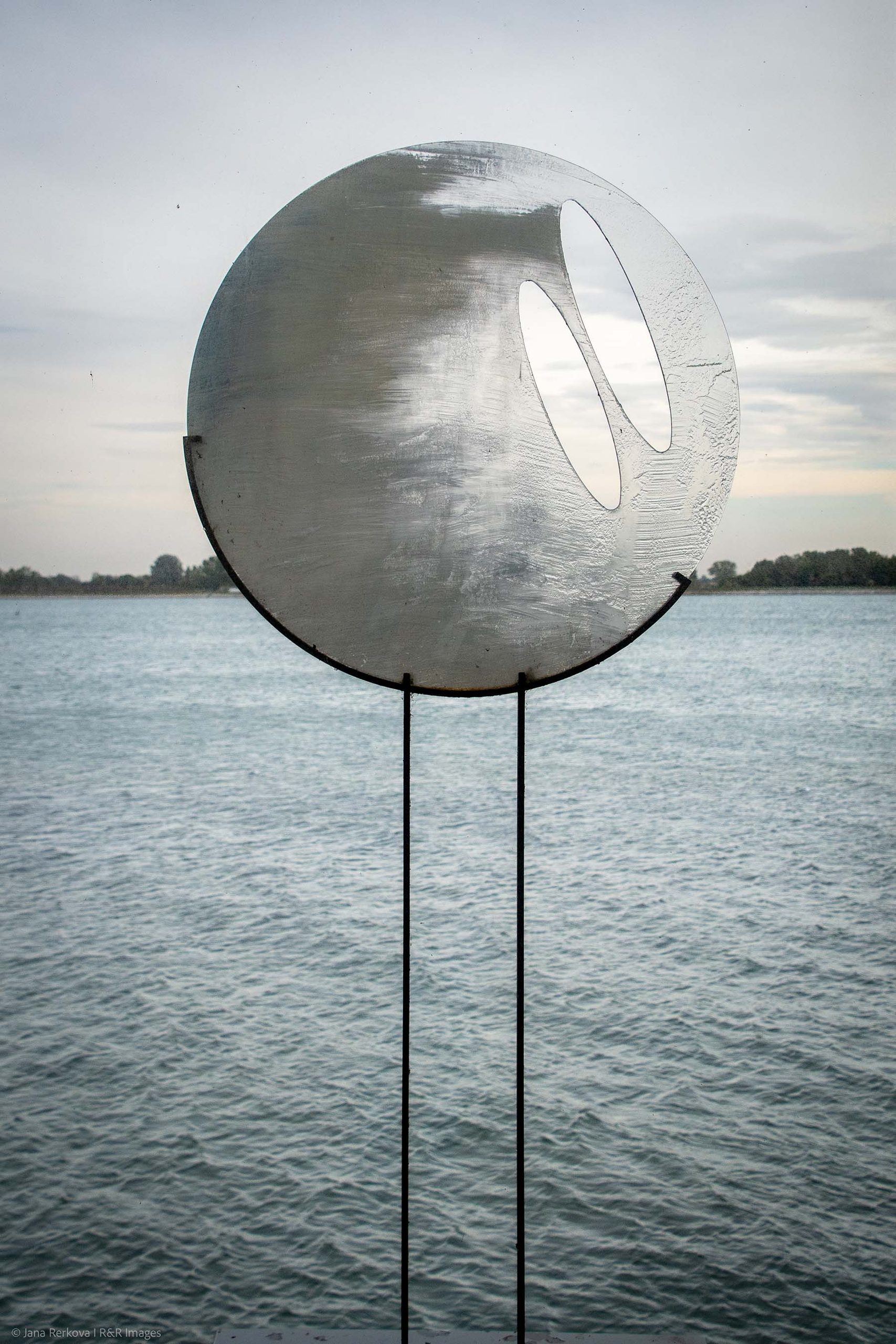 How Are You, Wind? by Palo Macho - A large metallic circular sculpture with two oval cutouts overlooking a wide river under a cloudy sky.